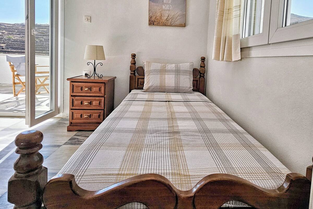 Een enkel bed met houten hoofdbord, een nachtkastje met lamp, een aan de muur hangend schilderij, en uitzicht door een glazen deur.