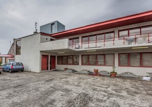 A two-story building with a red roof and a flat facade. There are several windows with red frames, and a car is parked in front.