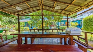 A covered outdoor dining area with wooden benches and a table, surrounded by lush greenery and a view of a blue building in the background. - Rat Villa (Pa Klok)