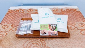 A wooden tray with a patterned tablecloth, containing white shower caps, a plastic container with cotton swabs, and a business card for a massage and spa service. - Rat Villa (Pa Klok)