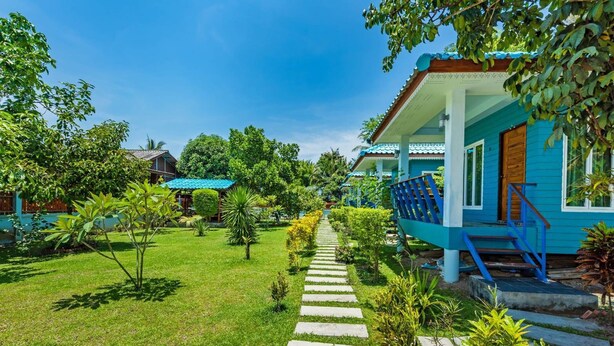 A blue building with a staircase leading to a porch, surrounded by a well-maintained garden with various plants and a clear sky. - Rat Villa (Pa Klok)