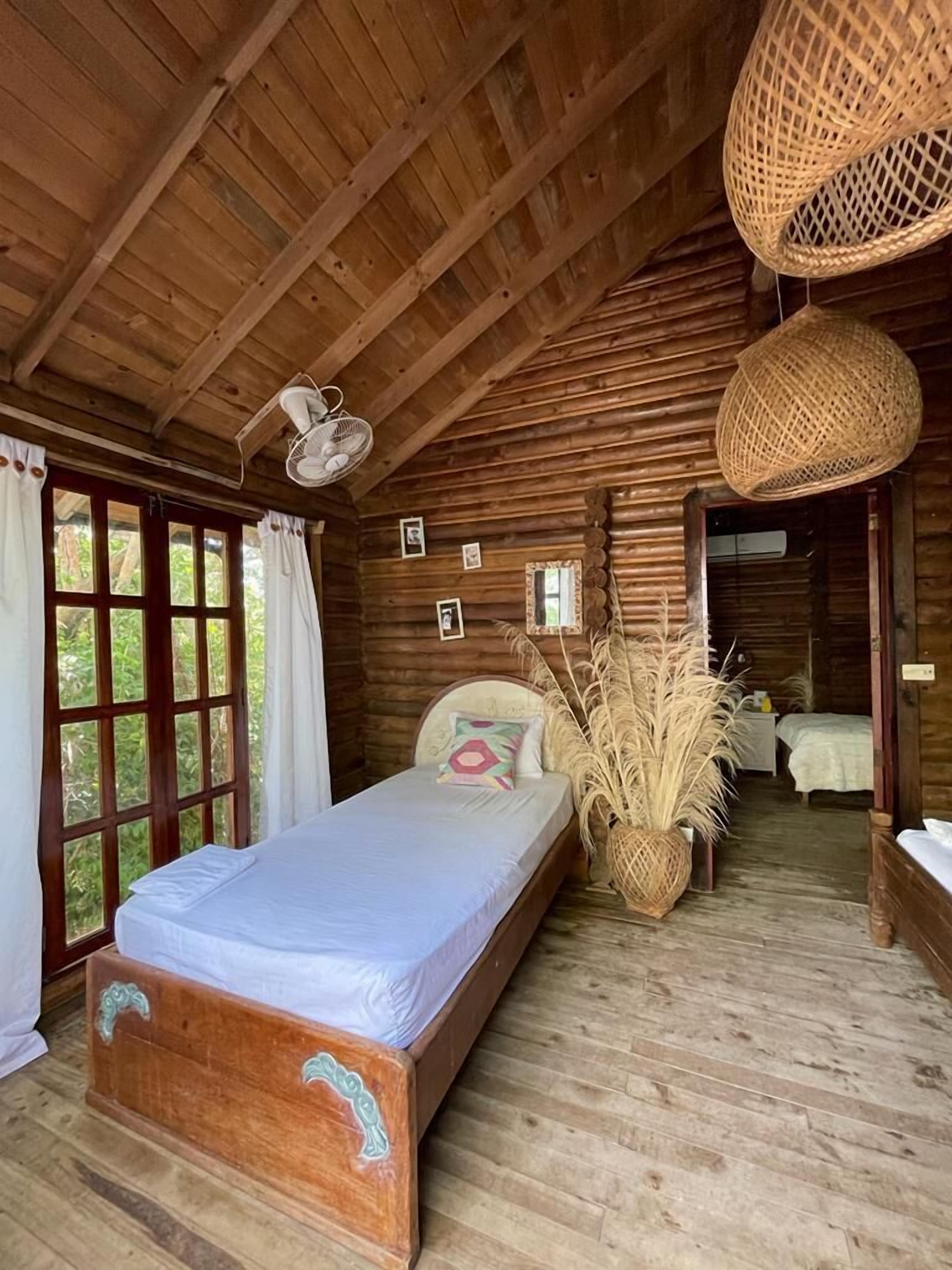 Un dormitorio de cabaña de madera con una cama individual, un ventilador colgante y un ventanal grande.