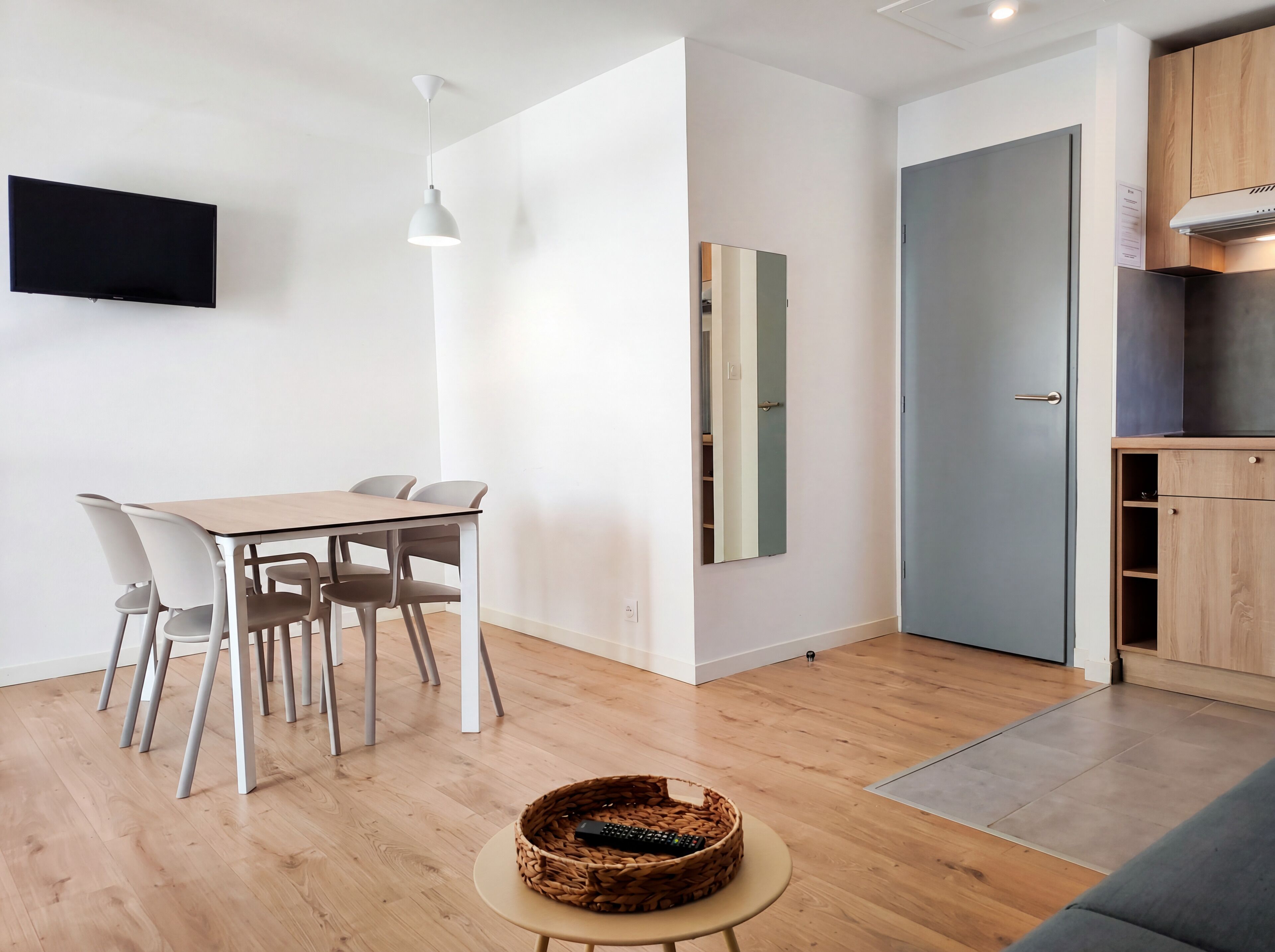 A modern room with a dining table, chairs, a TV, a mirror, and a kitchen area.