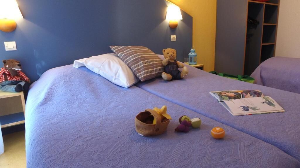 Una cama con un cojín a rayas, un peluche, una cesta de frutas y una revista.
