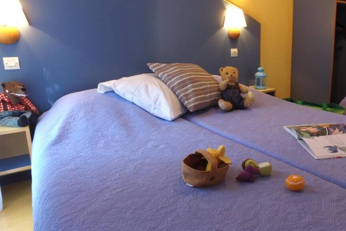 Un lit avec un oreiller Ă rayures, un ours en peluche, un panier de fruits et un magazine.