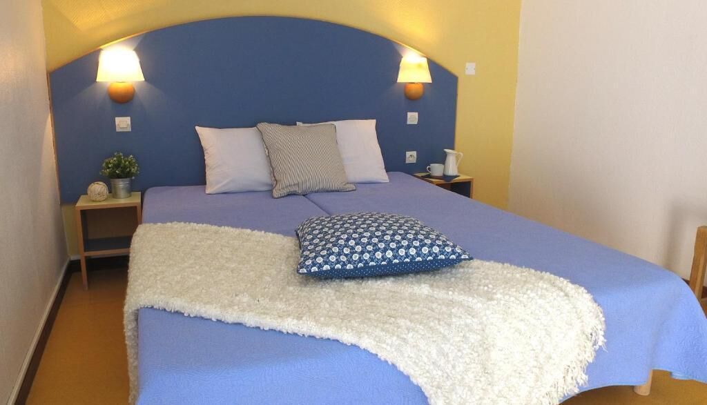 Una cama con cabecera azul, dos lámparas de noche, una manta blanca y un cojín estampado.