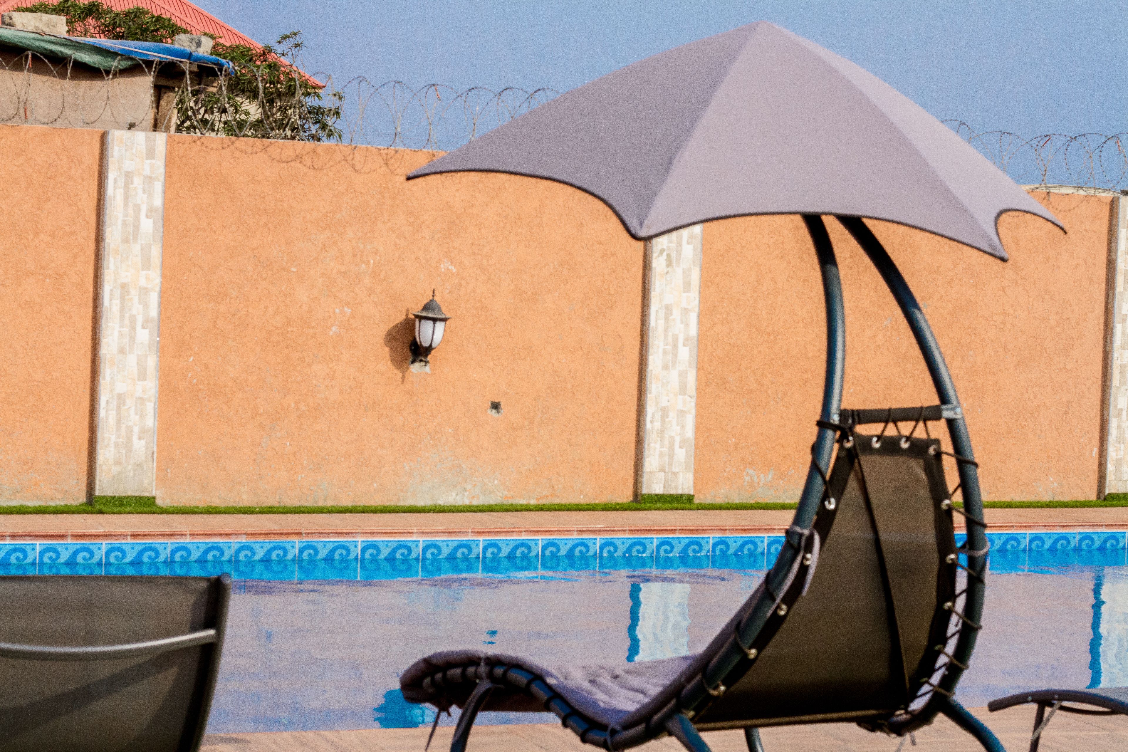 Zona junto a la piscina con una silla de descanso bajo un paraguas grande, una piscina con baldosas y una pared con alambre de púas.