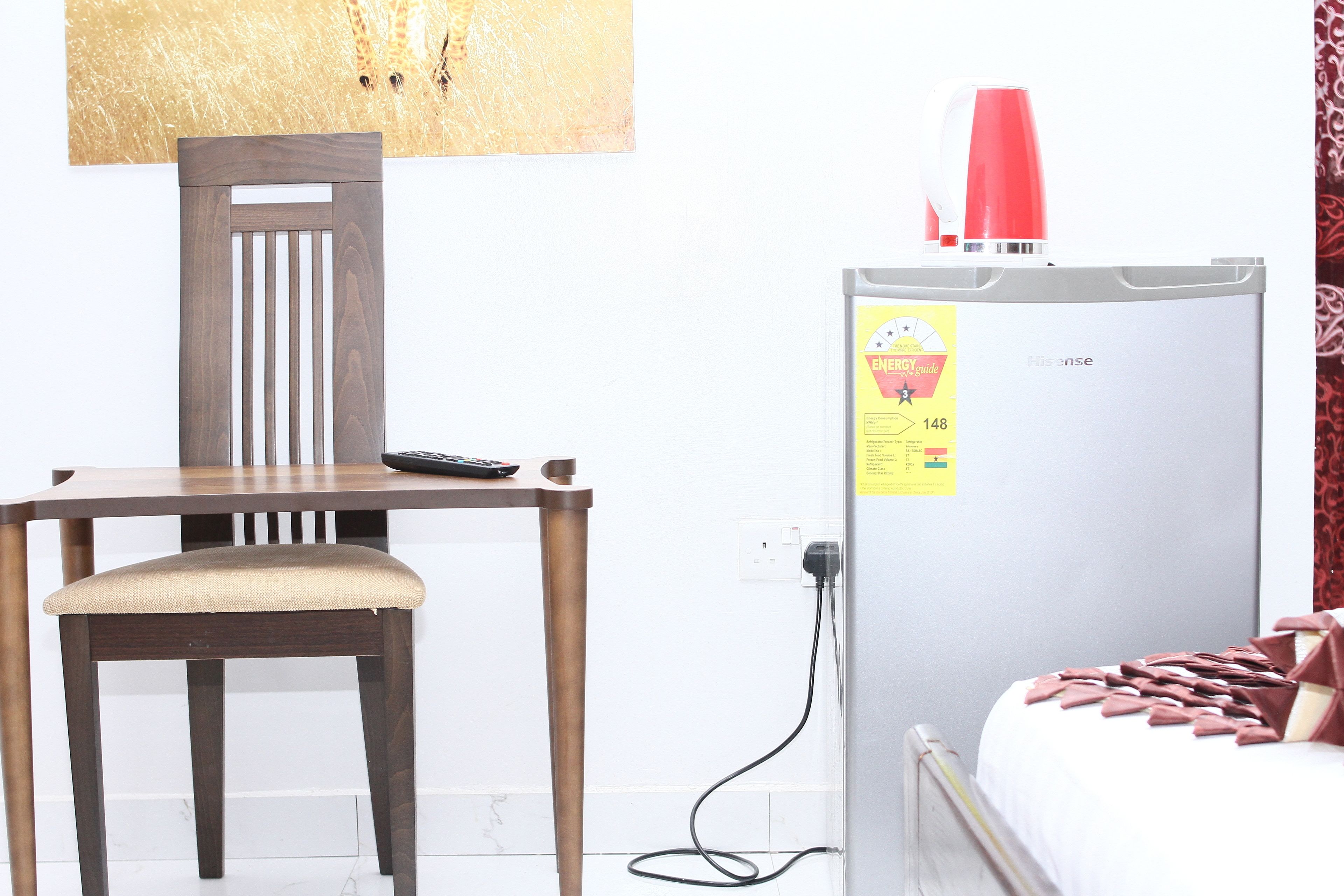 A room with a wooden chair, a white bed with a red blanket, a white refrigerator with a red lamp on top, and a wall-mounted power outlet.