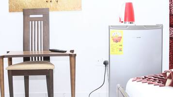 A room with a wooden chair, a white bed with a red blanket, a white refrigerator with a red lamp on top, and a wall-mounted power outlet.