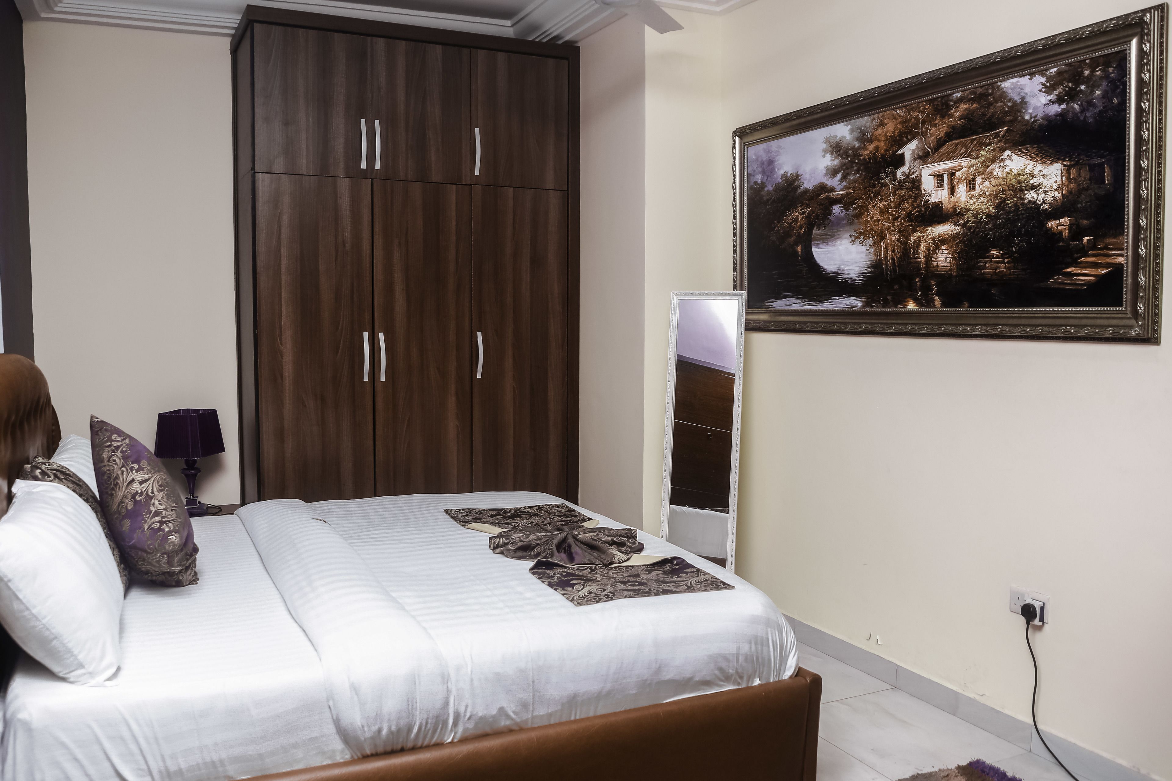 A bedroom with a bed, a wardrobe, a painting, and a bedside table with a lamp.