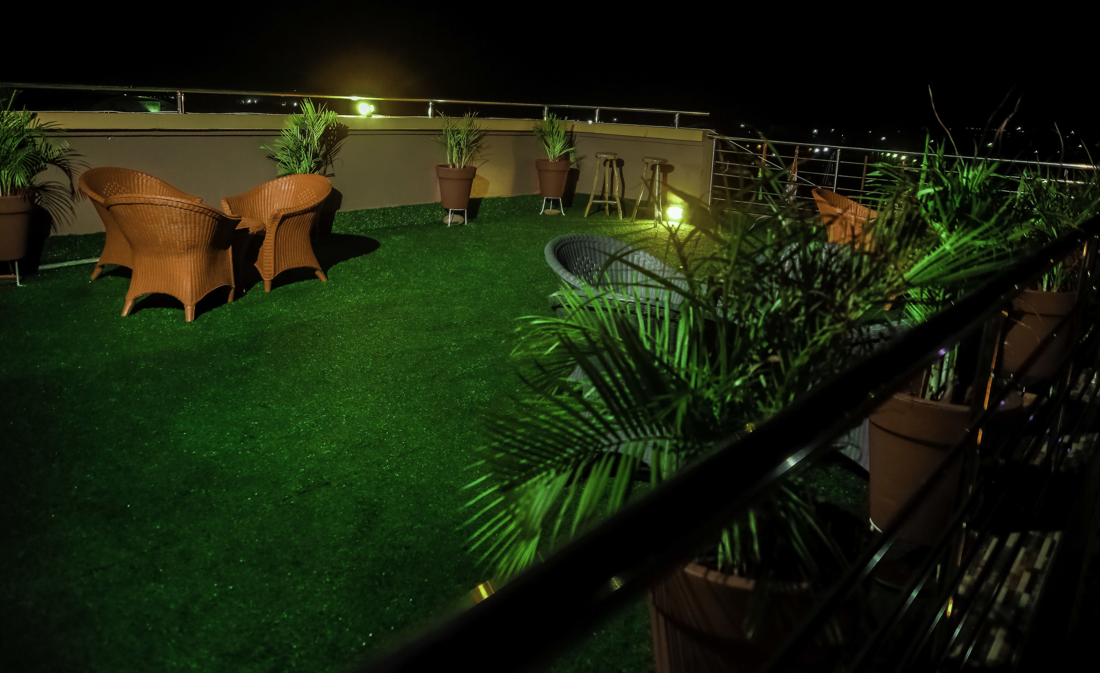 A rooftop terrace with artificial grass, wicker chairs, potted plants, and a metal railing.