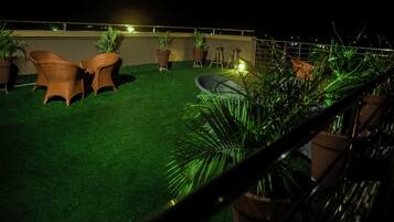 A rooftop terrace with artificial grass, wicker chairs, potted plants, and a metal railing.