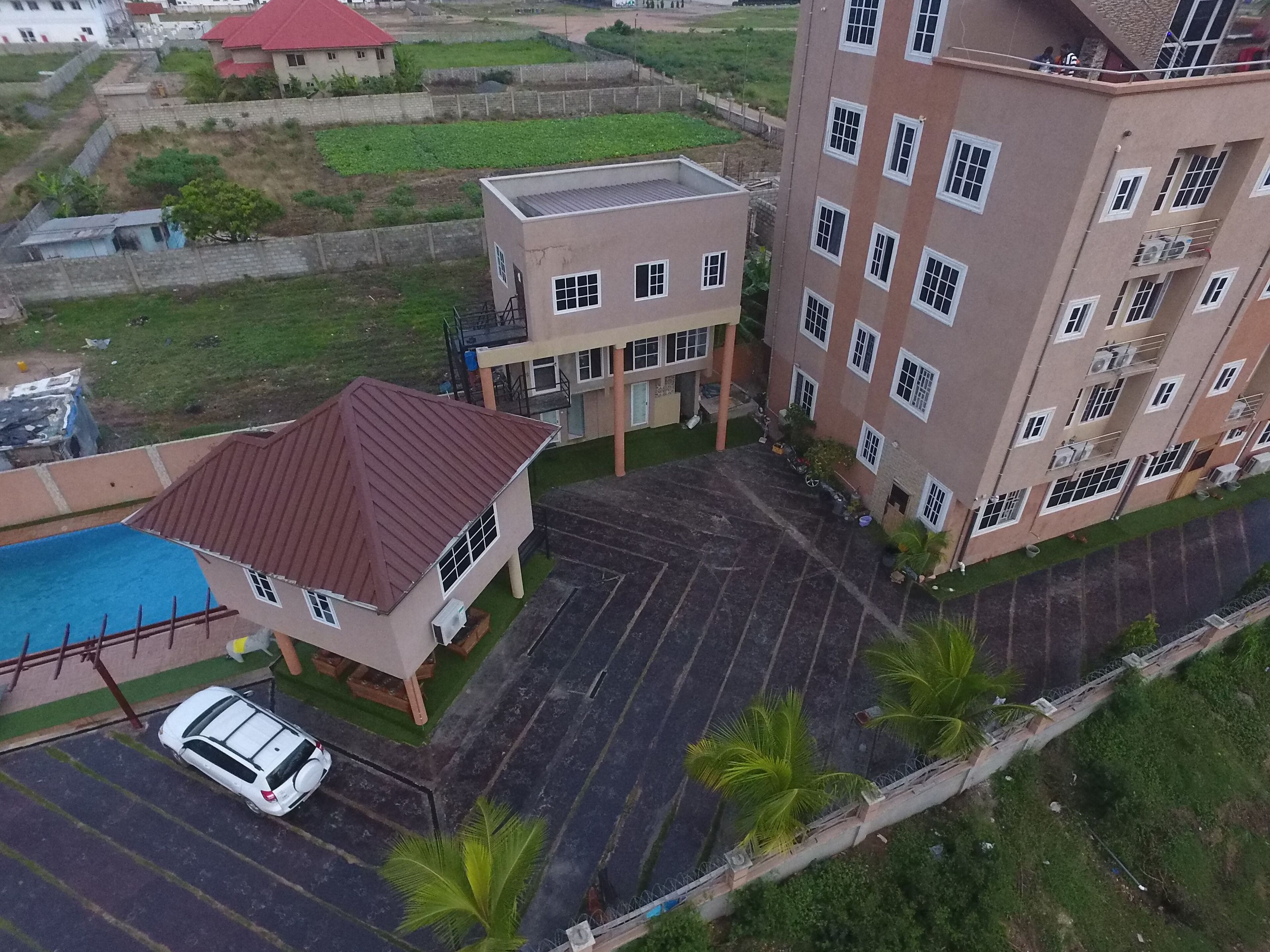 Vista aérea de un edificio residencial de varios pisos, con un área de estacionamiento, piscina y un edificio pequeño adyacente.