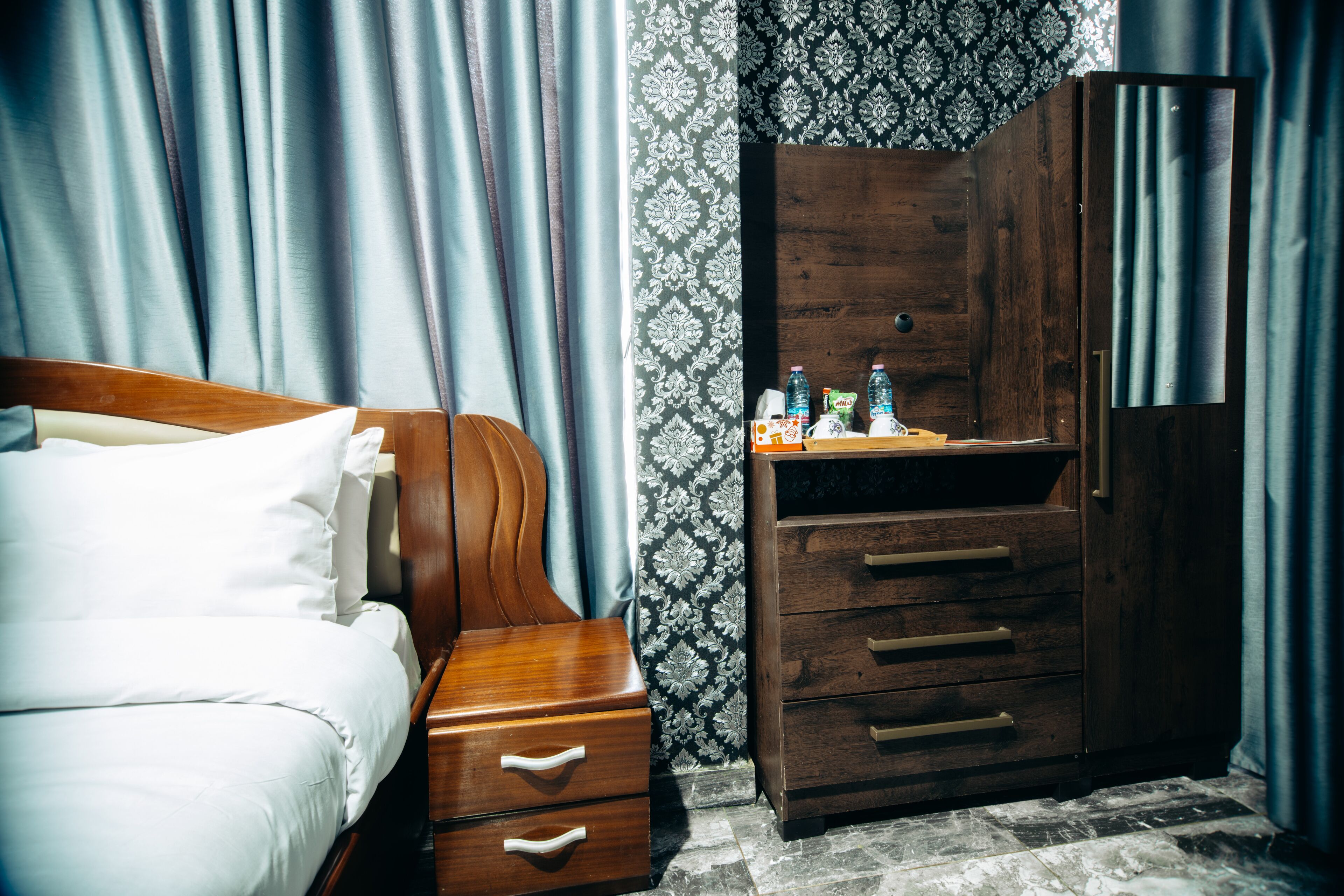 Een hotelkamer met een houten bed, nachtkastje, dressoir met lades en een spiegel.