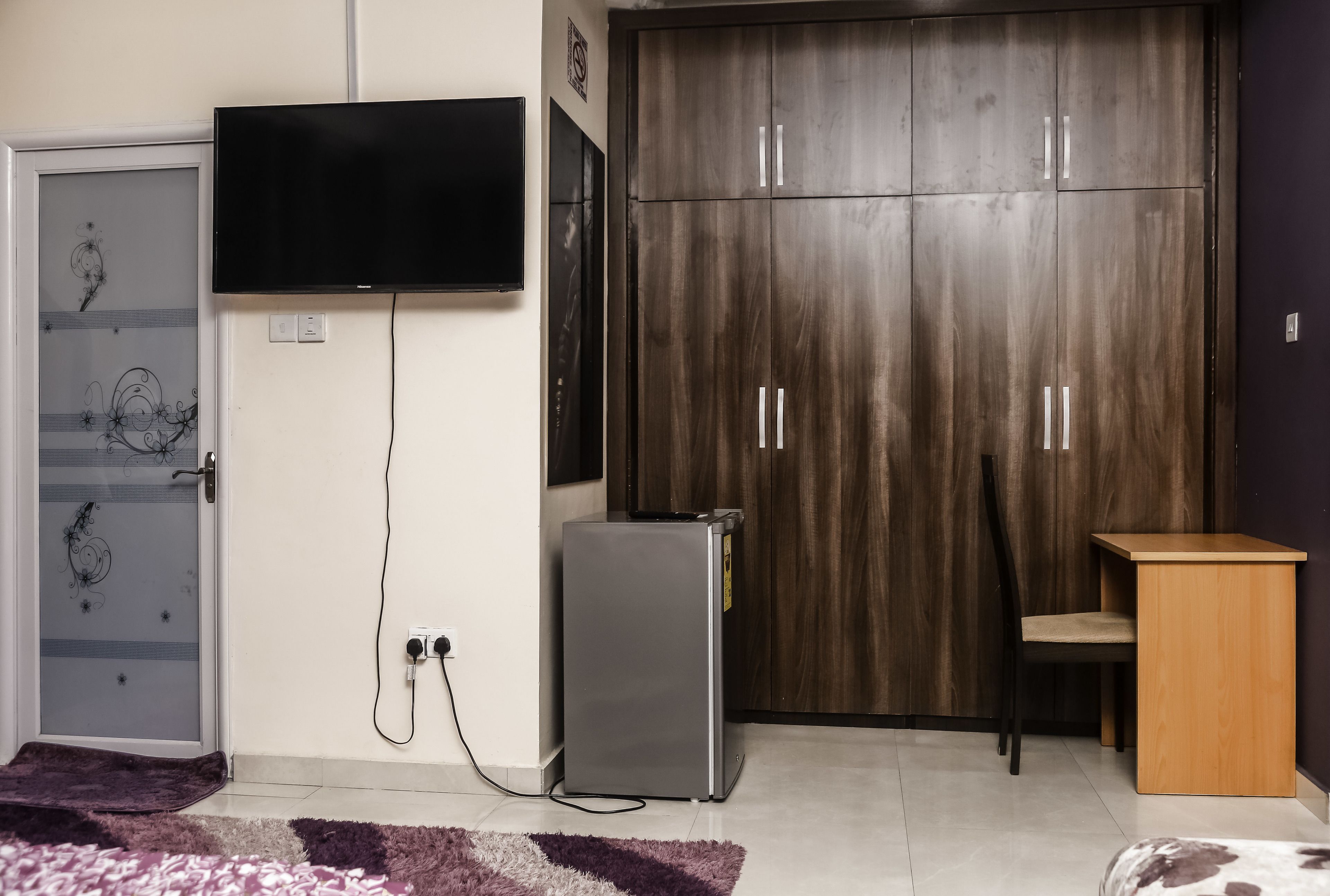Una habitación con televisión montada en la pared, armario, refrigerador pequeño, escritorio y una silla.