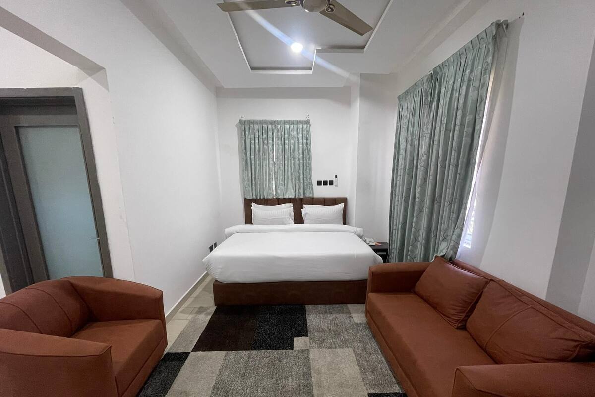 Habitación de hotel con cama, dos sofás, ventilador de techo y alfombra estampada.