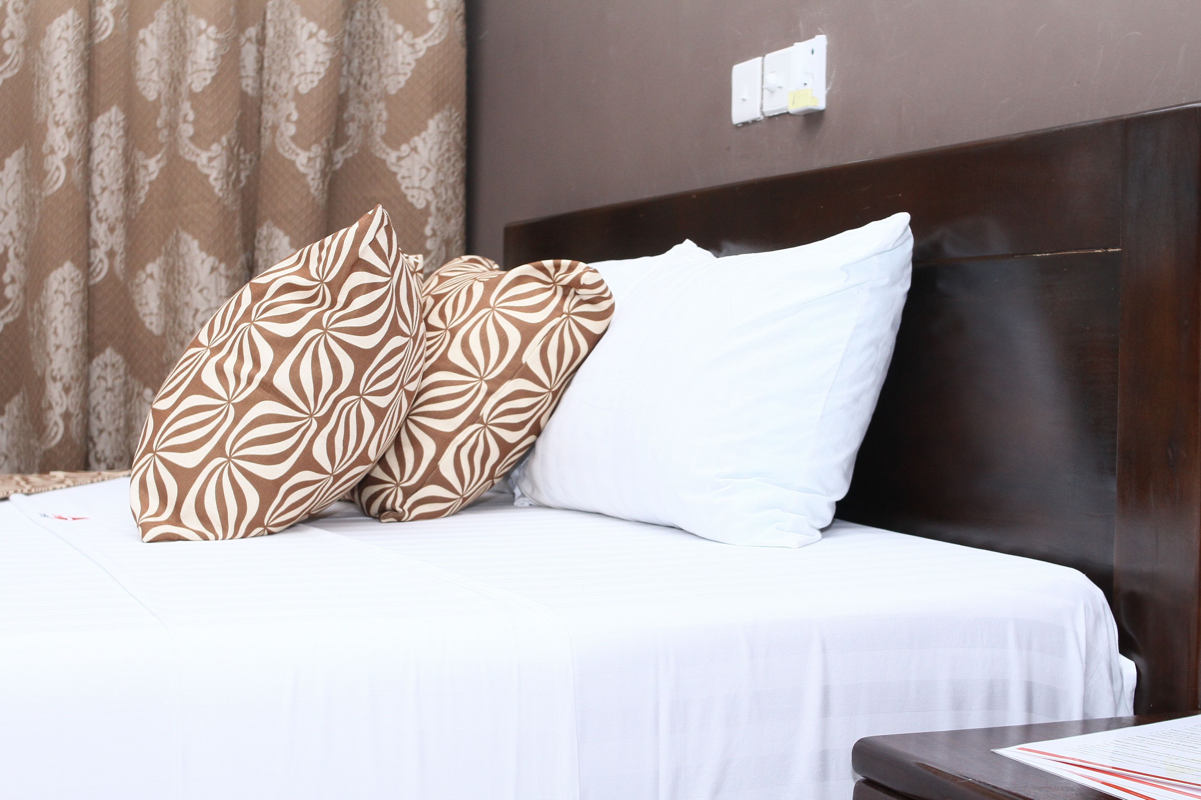 Una cama bien tendida con ropa blanca y almohadas estampadas, un cabecero oscuro y una pared marrón con un enchufe.