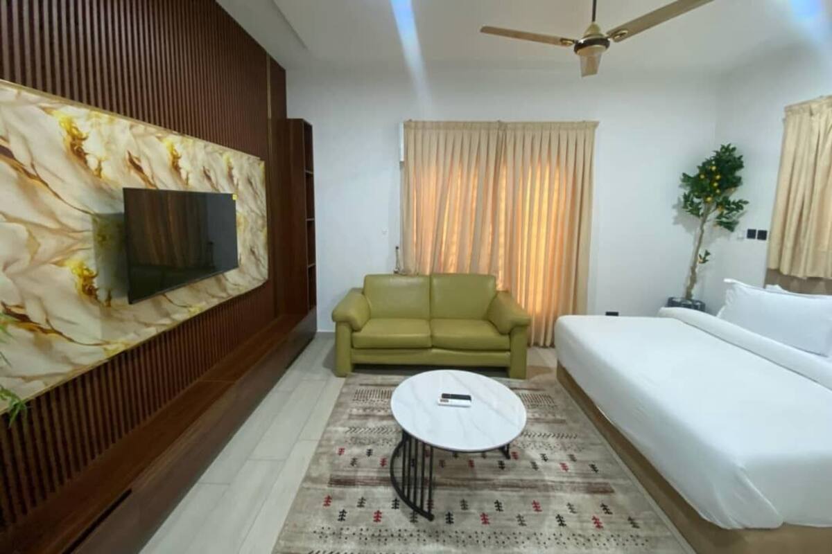 Una habitación de hotel moderna con un televisor de pantalla plana montado en un panel de madera, un sofá verde, una mesa de centro redonda, una cama con ropa de cama blanca y un ventilador de techo.