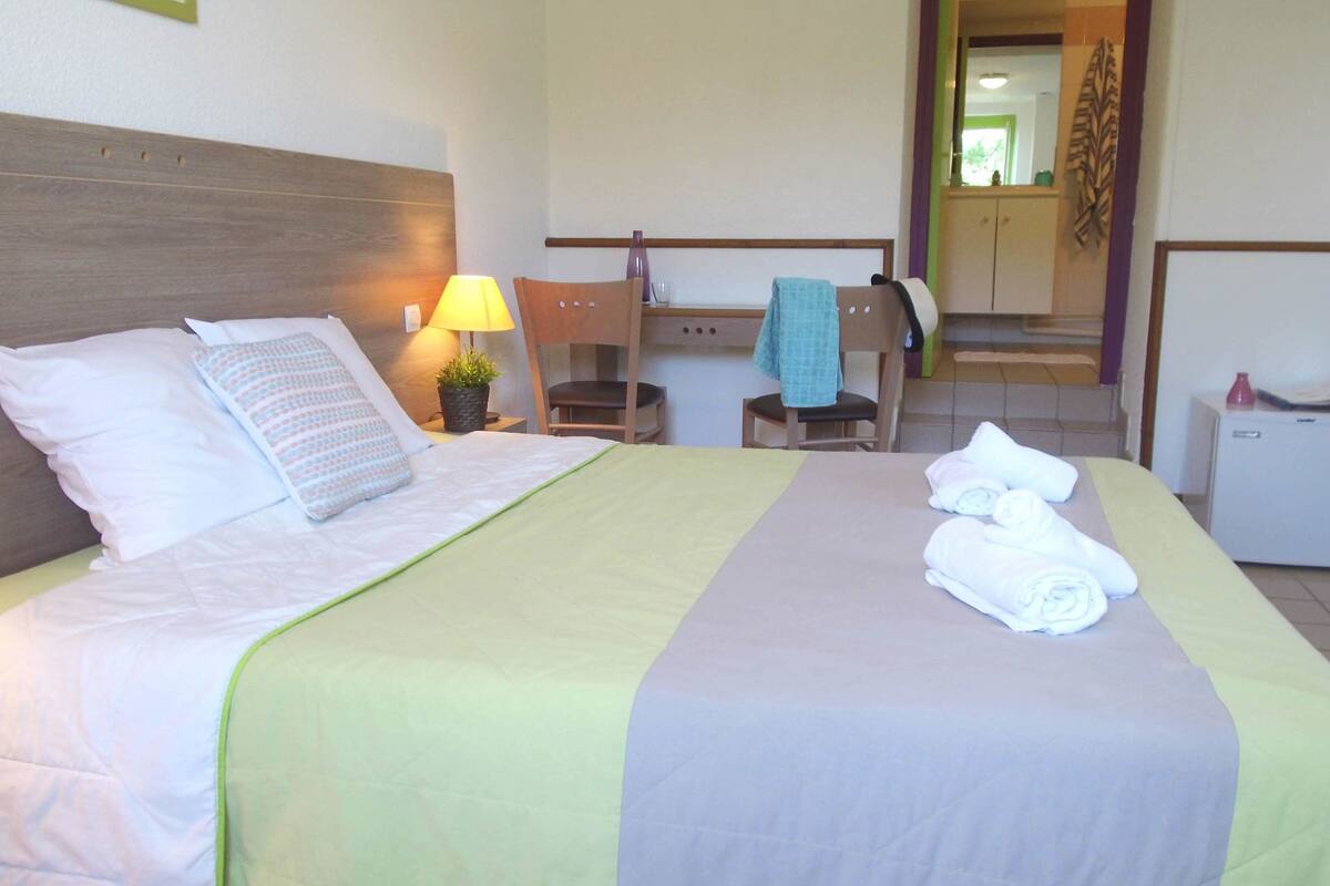 Un lit bien fait, avec des oreillers et une couverture, une lampe de chevet, une chaise, une petite table avec une serviette et un réfrigérateur.
