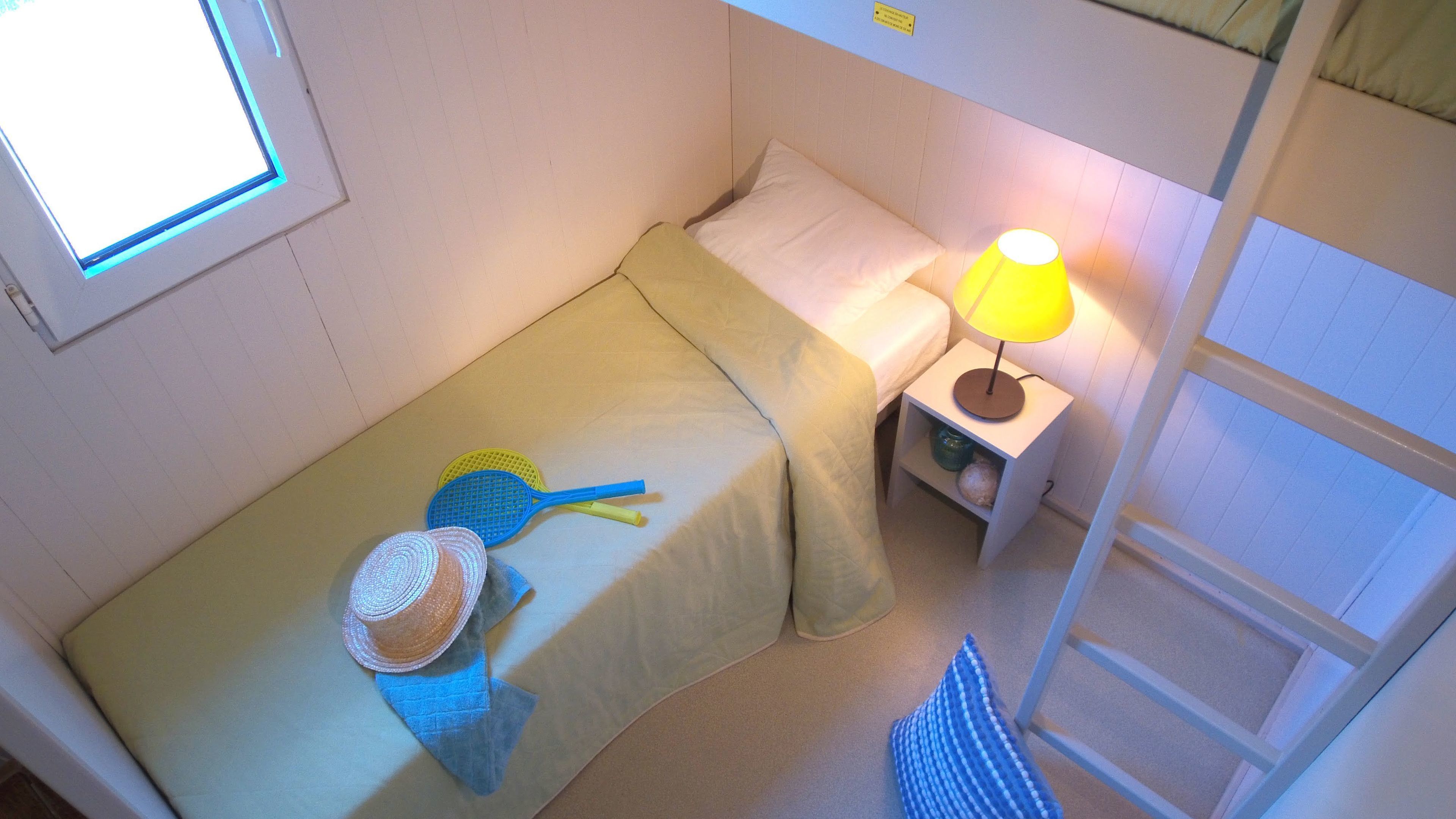 A small, single-bed room with a window, a bedside lamp, and a small shelf with a lamp and a vase.