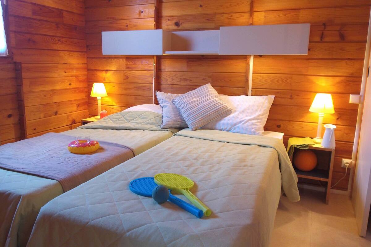Une chambre dans un chalet en bois, avec deux lits, chacun muni d’un oreiller et d’une lampe. On y trouve une petite table sur laquelle sont posés une lampe et une bouteille.