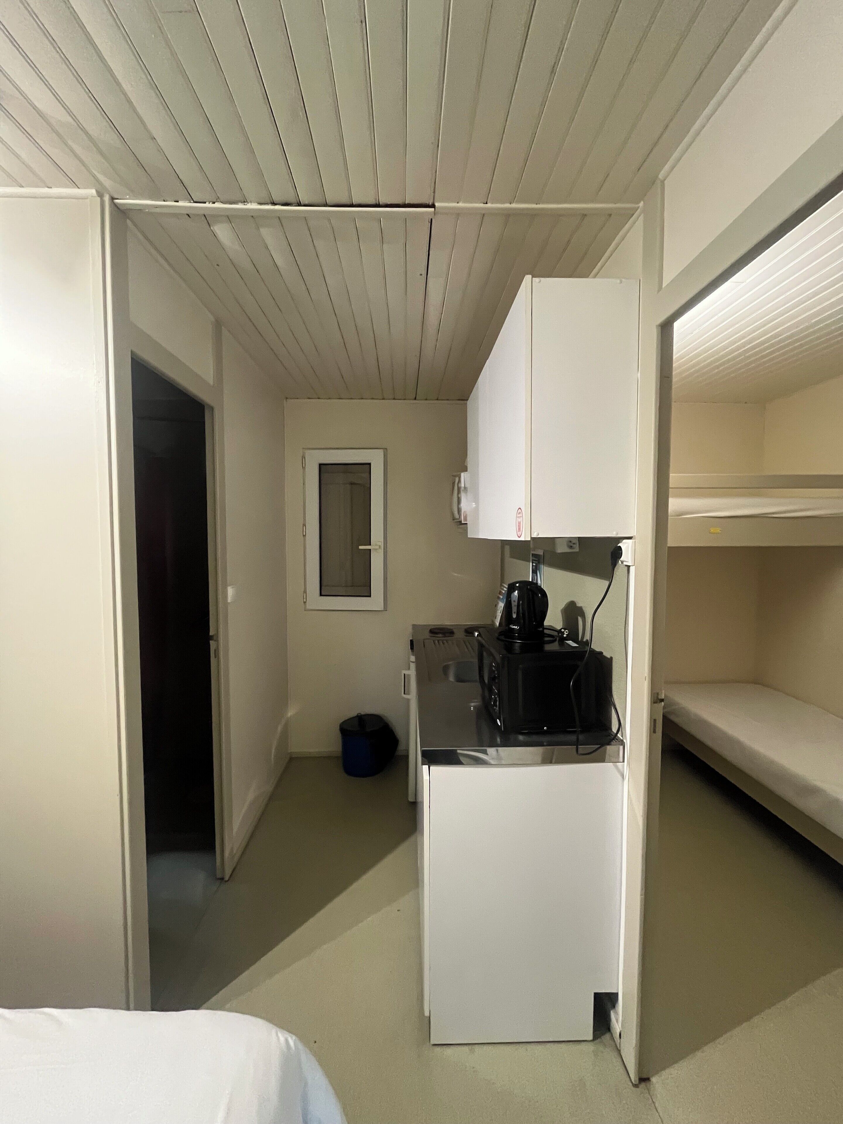 Une chambre d’hôtel de taille réduite, dotée d’une kitchenette, d’un lit simple et d’une petite fenêtre.