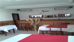 Un restaurant dont les tables sont recouvertes de nappes blanches et de chemins de table rouges, avec des chaises en bois, un arrangement floral central et un bar où les bouteilles sont exposées sur des étagères.
