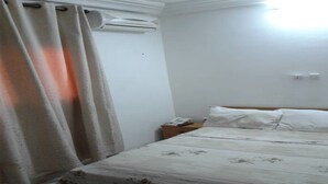 Une chambre à coucher comprenant un lit, des rideaux, un appareil de climatisation, une table de chevet avec un téléphone et une petite table de nuit.