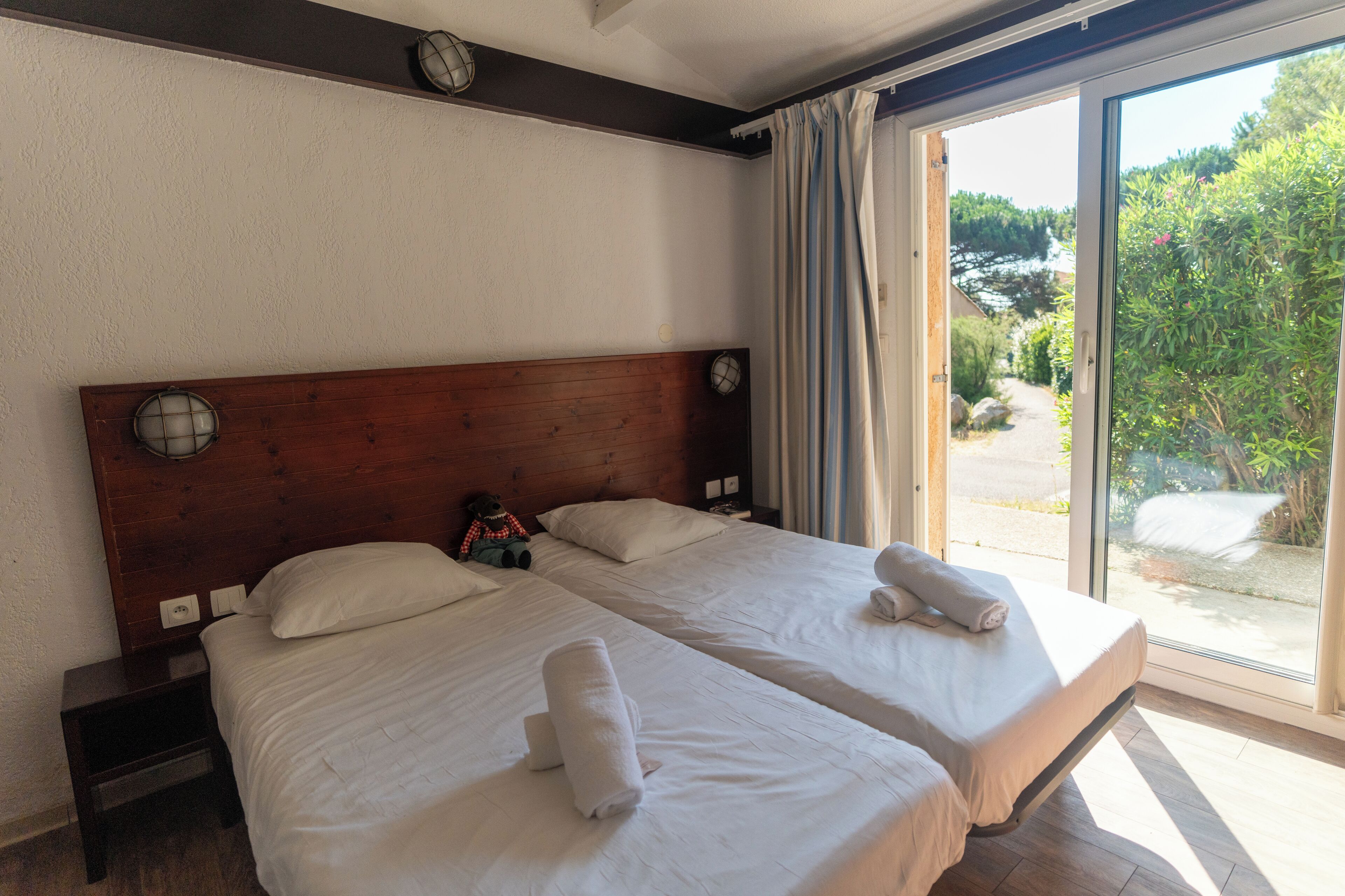 Een tweepersoonskamer met twee bedden, een houten hoofdbord, een nachtkastje, een klok en uitzicht op groen via een schuifdeur.