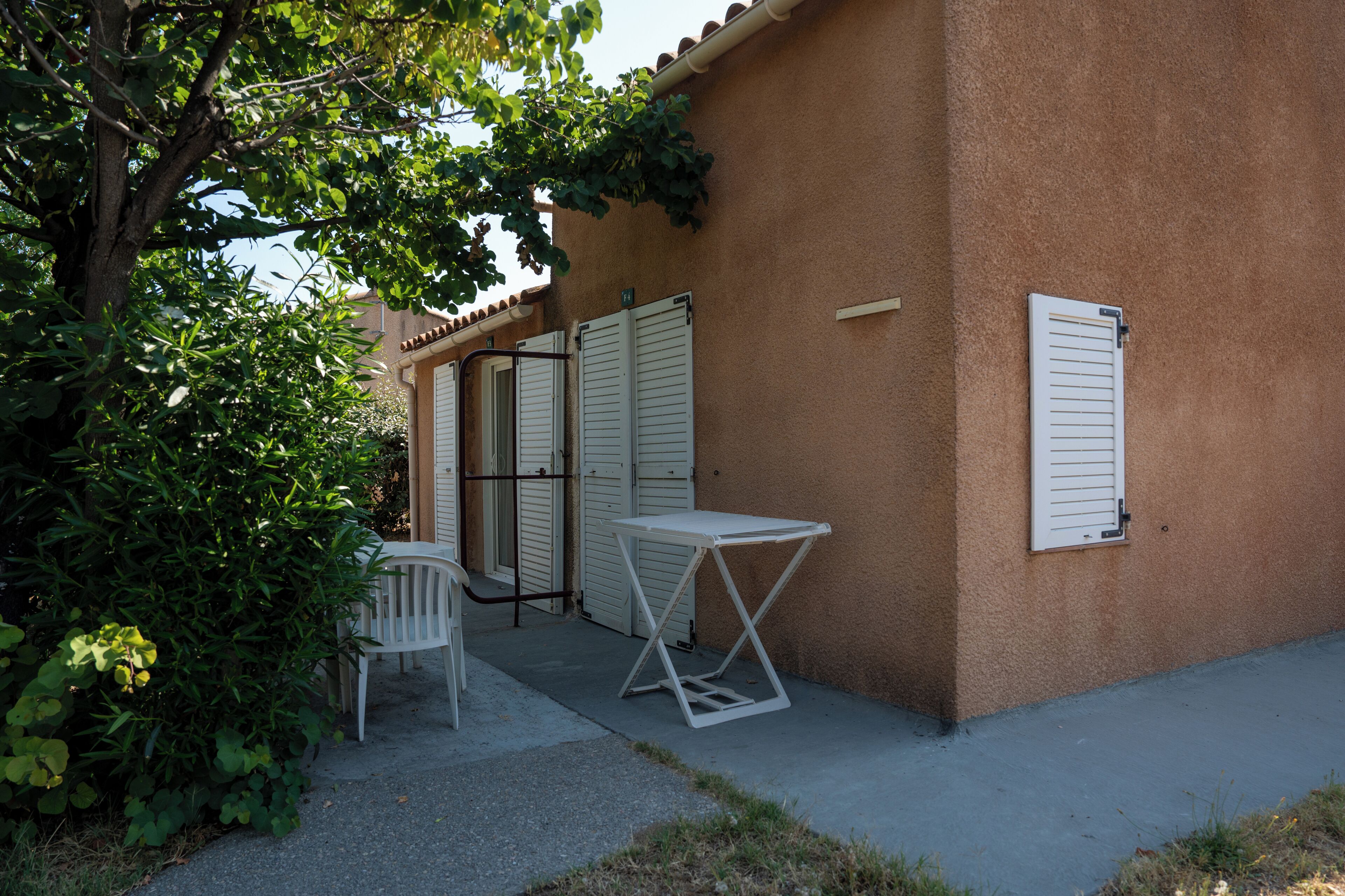 A small outdoor seating area with a table and chairs, a tree, and a building with shutters.