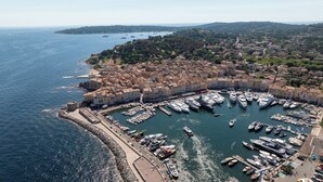 An aerial view of a coastal town with a marina filled with boats, surrounded by historic buildings and a hilly landscape in the background.