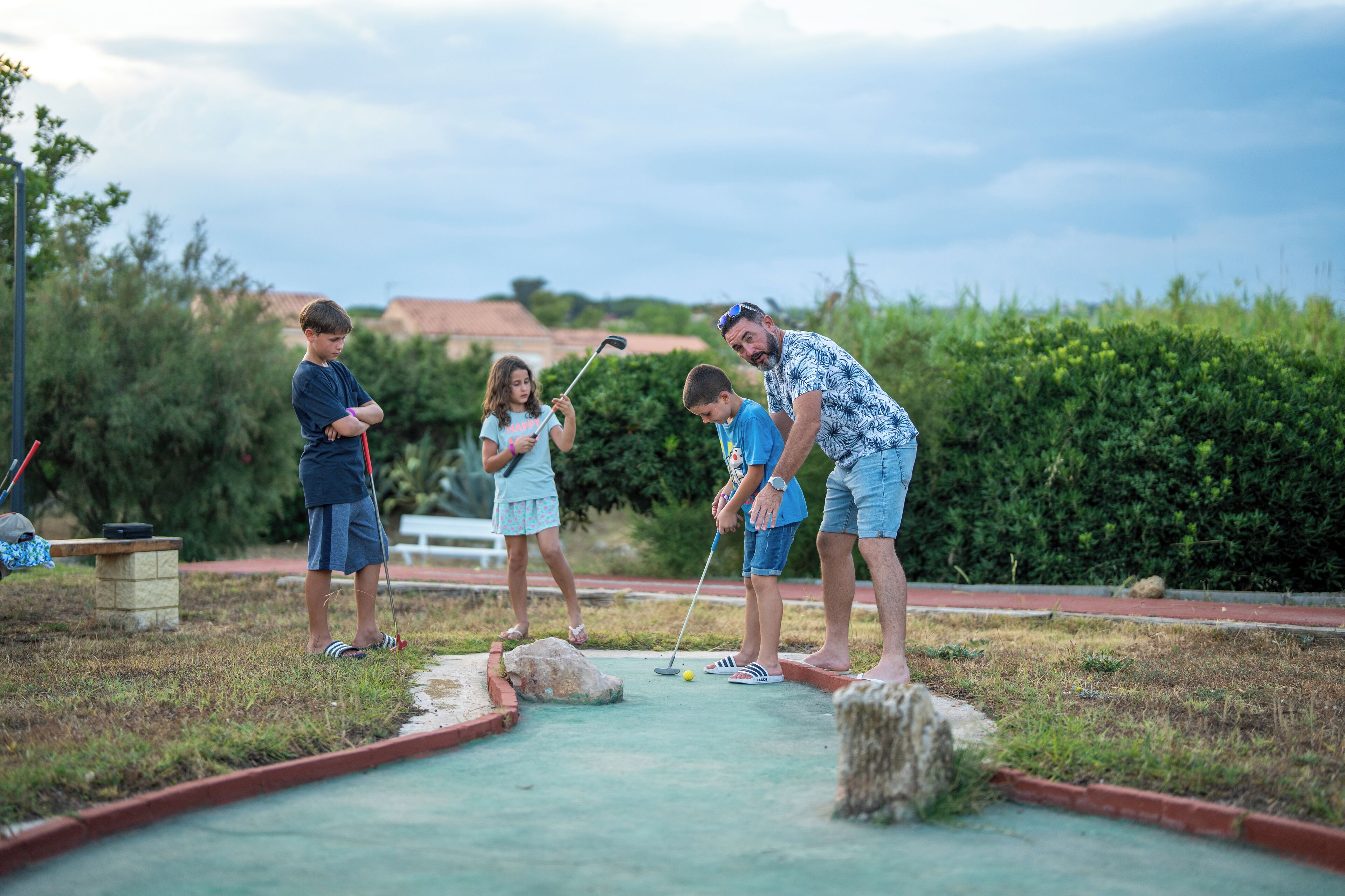 A group of people playing croquet on a grass field with a small pond and trees in the background.