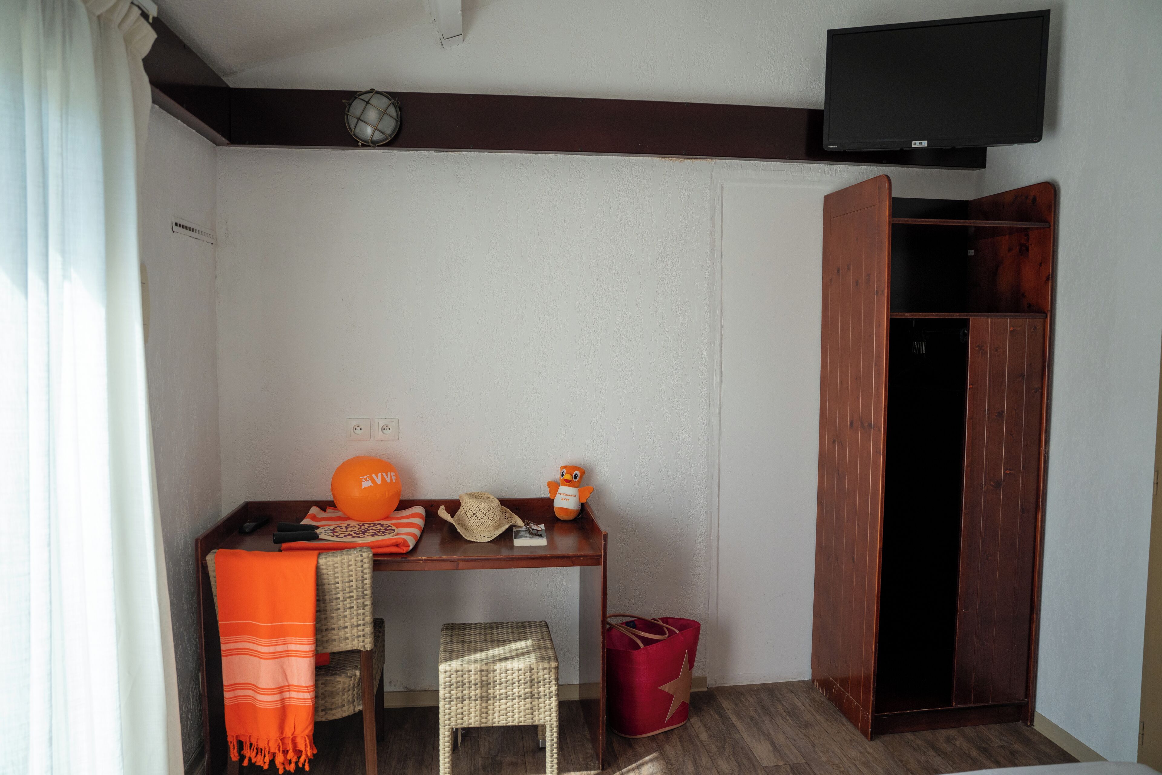 Une pièce comprenant un bureau en bois, une chaise en osier, un sac rouge, un téléviseur fixé au mur et une armoire en bois.