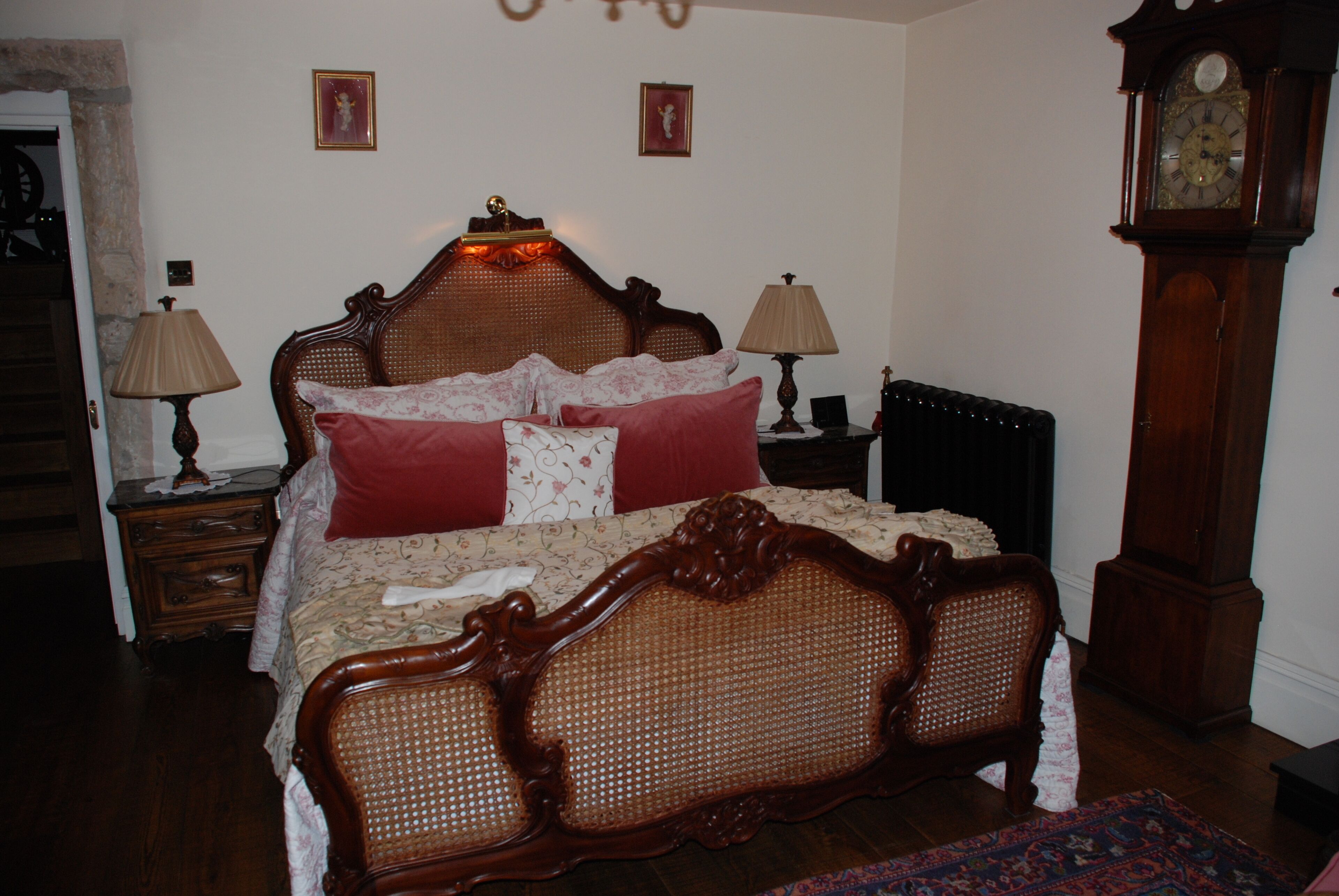 Une chambre à coucher de style traditionnel, avec un lit à baldaquin en bois, deux tables de chevet avec des lampes, une grande horloge murale et un radiateur.