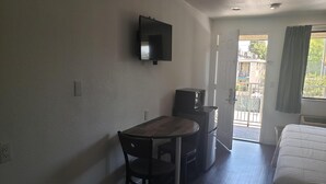 A compact room with a bed, a small table, a chair, a TV mounted on the wall, and a door leading to a balcony.