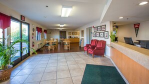 A hotel lobby with a reception desk, red chairs, a dining area with tables and chairs, and a large potted plant.