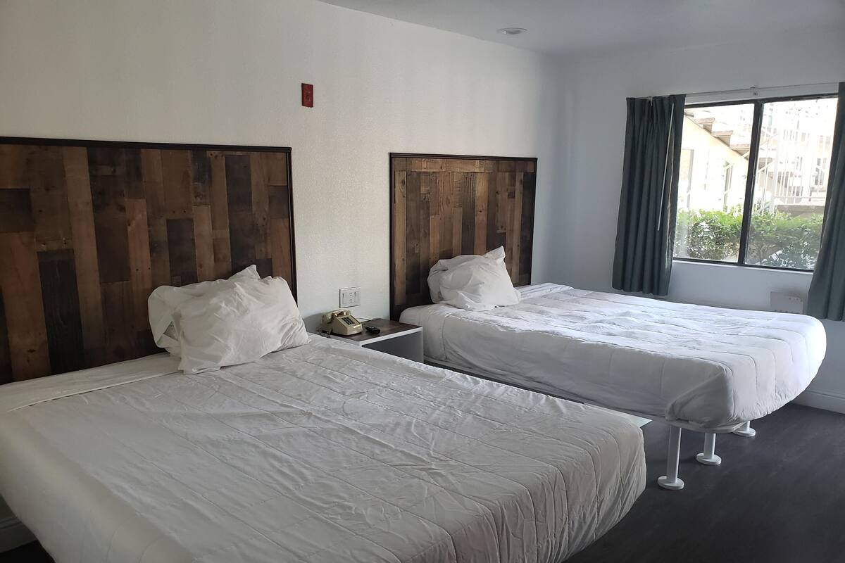 Habitación de hotel con dos camas, cabeceros de madera, ropa de cama blanca, un teléfono en la mesita de noche y una ventana con cortinas.