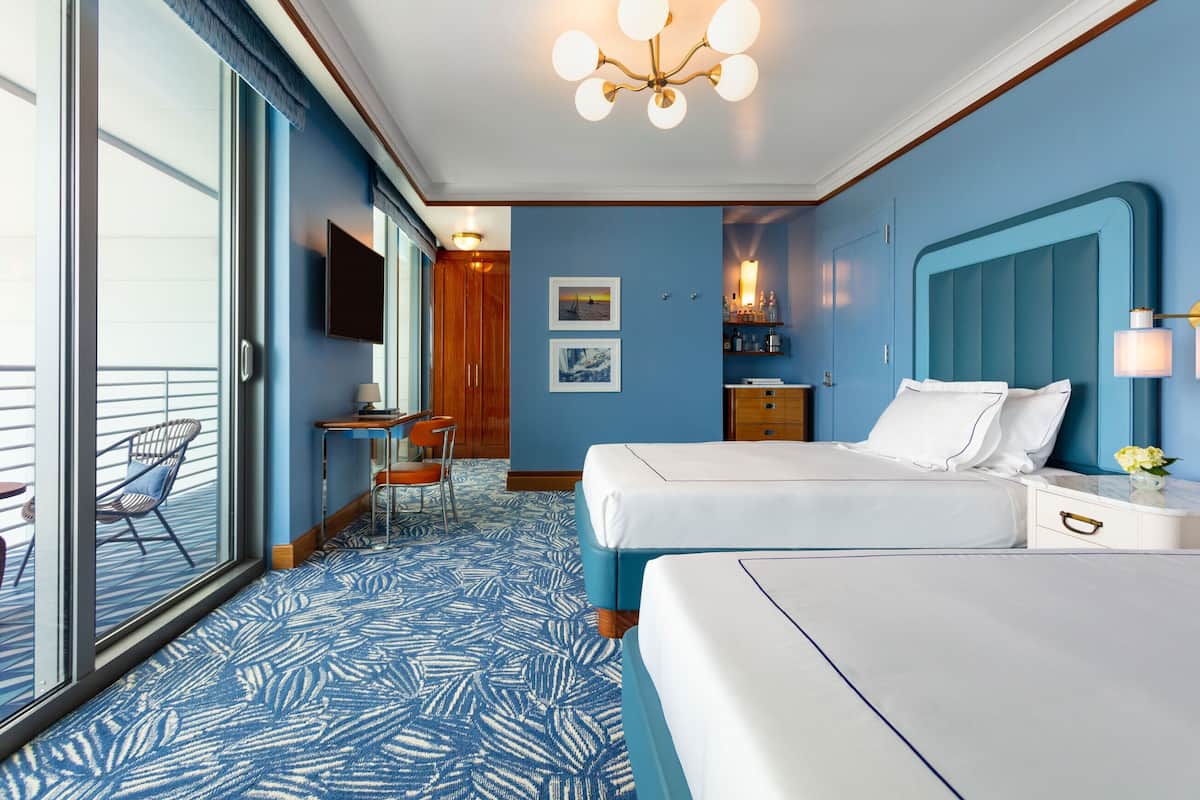 A hotel room with two beds, a desk, a chair, a TV, and a balcony with a view.