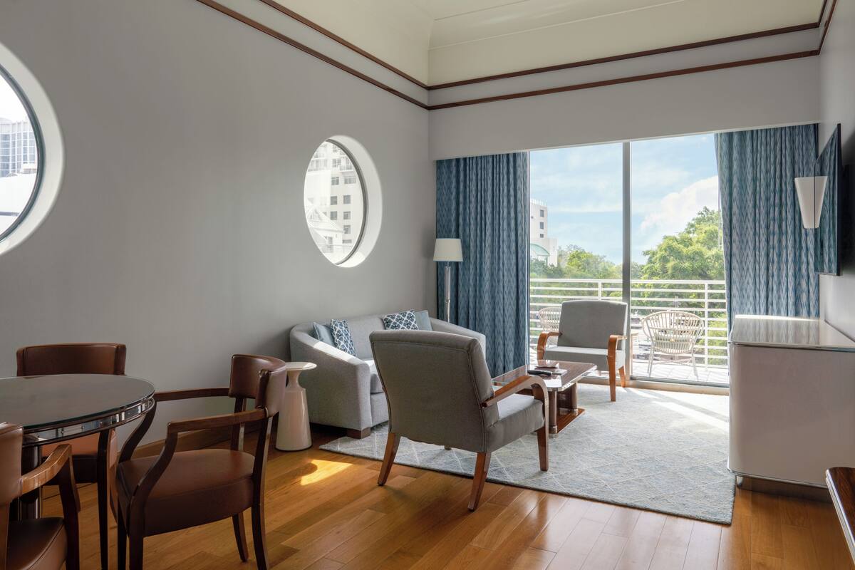 A modern hotel room with a round table, two sofas, a TV, and a balcony with a view.