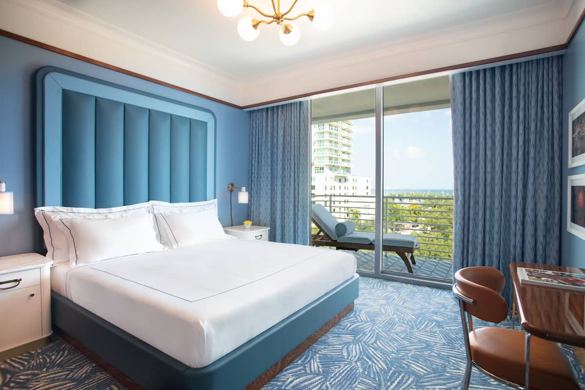 A hotel room with a large bed, a desk with a chair, a balcony with a view of the city, and a large window with curtains.