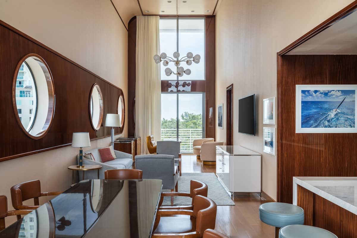 A modern hotel room with a large window, a dining area, and a seating area with a sofa and chairs.