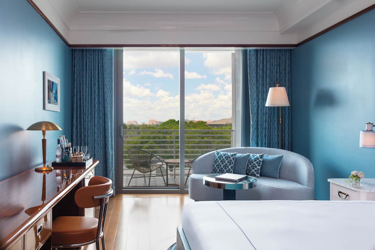 A modern hotel room with a large bed, a seating area with a sofa and chairs, a desk with a lamp, and a view of the outdoors.