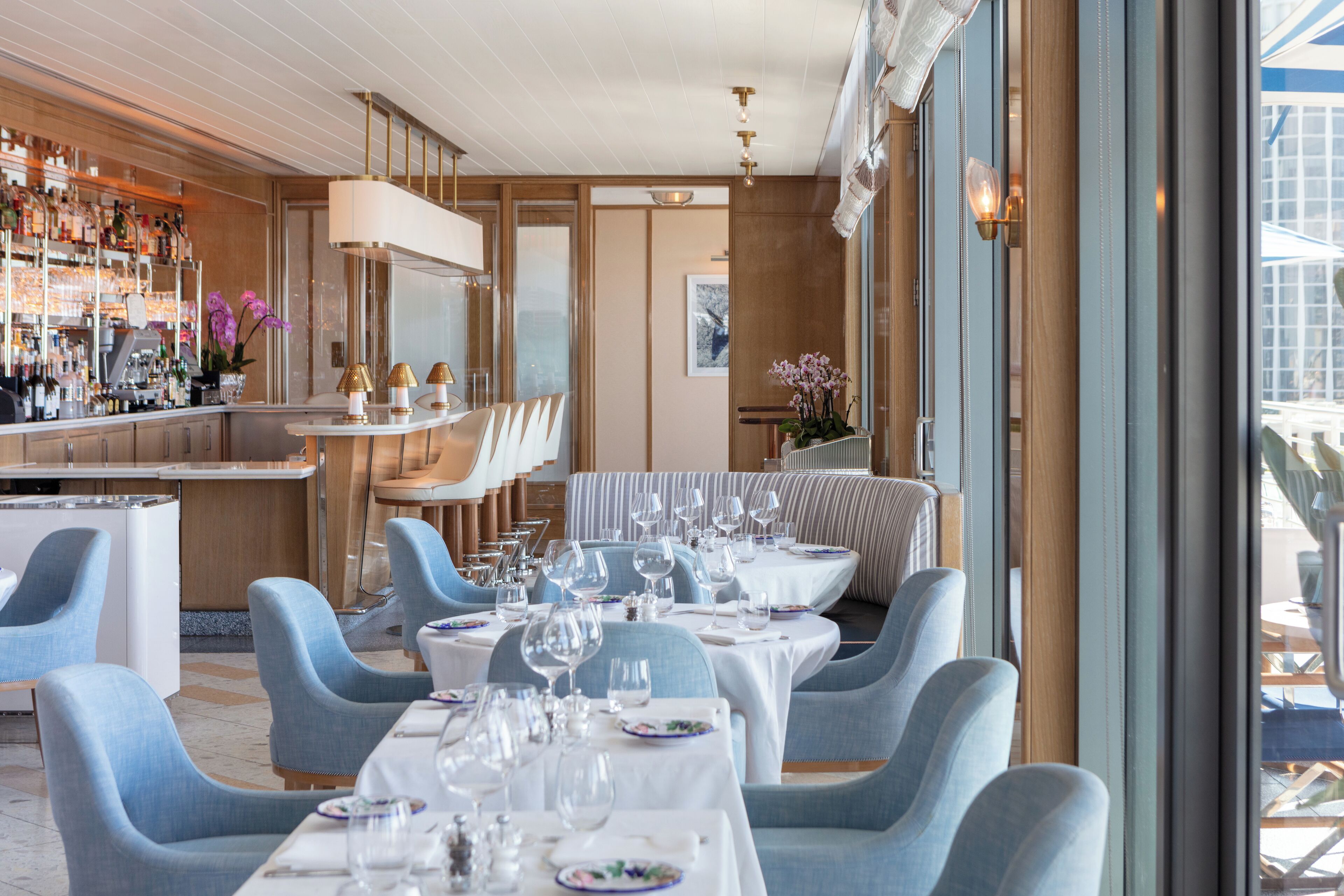A restaurant with round tables set for dining, blue upholstered chairs, and a bar area with shelves of bottles.
