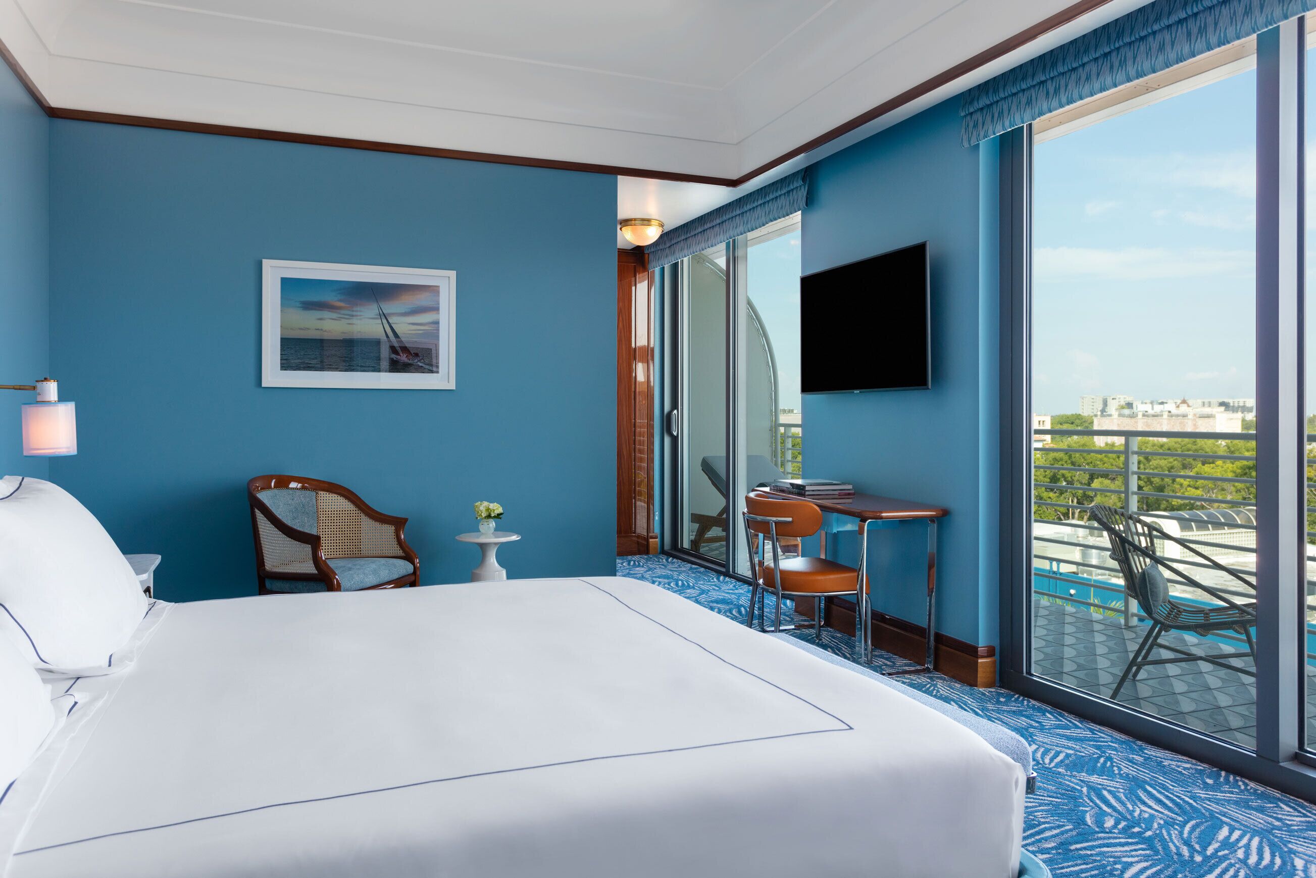 A hotel room with a large bed, a small table with chairs, a TV, and a balcony with a view of the city.