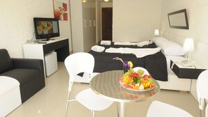 A hotel room with a bed, a desk with a computer, a TV, a dining table with fruit, and a white sofa. - Palmera Beach Hotel (Chihuahua)