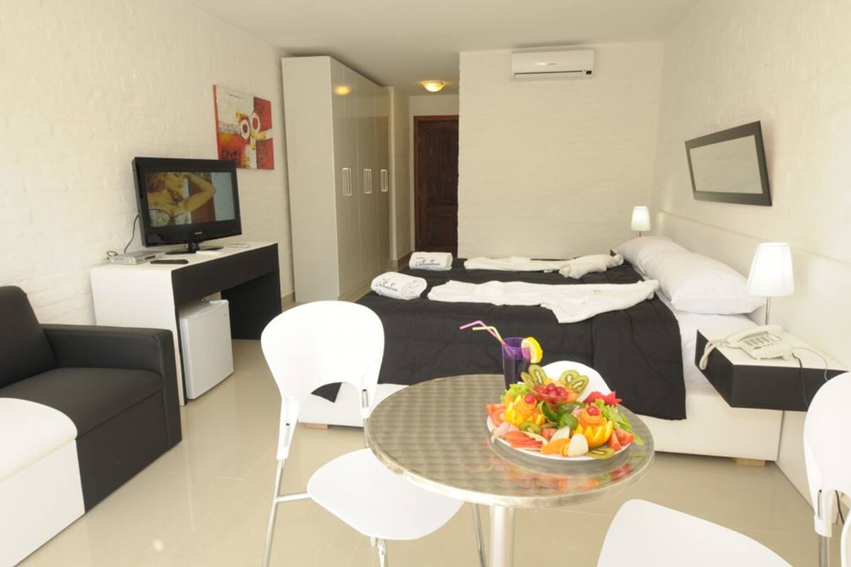 A hotel room with a bed, a desk with a computer, a TV, a dining table with fruit, and a white sofa.
