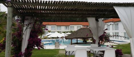A covered outdoor area with a pool, white tables, and chairs, surrounded by greenery and a building with a red roof.