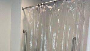 Un área de ducha con cortina transparente, barras de apoyo metálicas y pared de azulejos blancos.