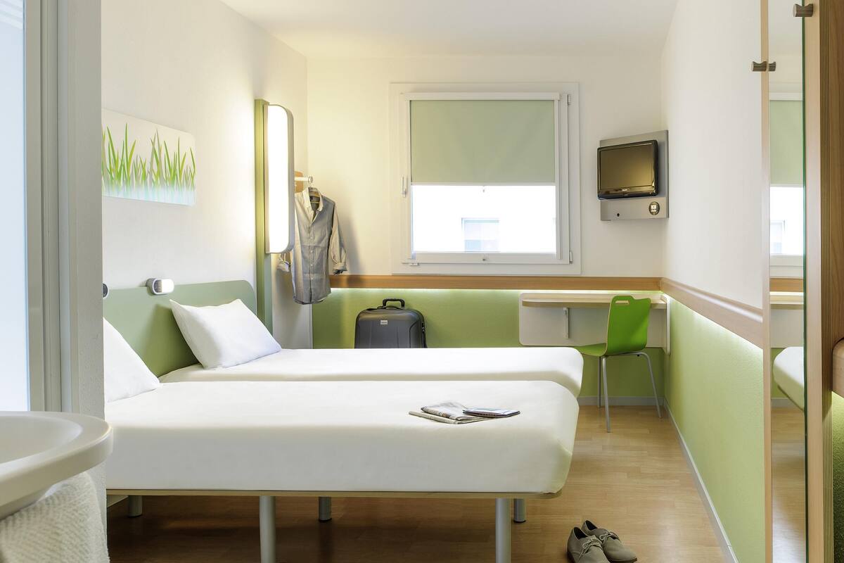 A hotel room with two beds, a desk, a chair, a suitcase, and a TV.