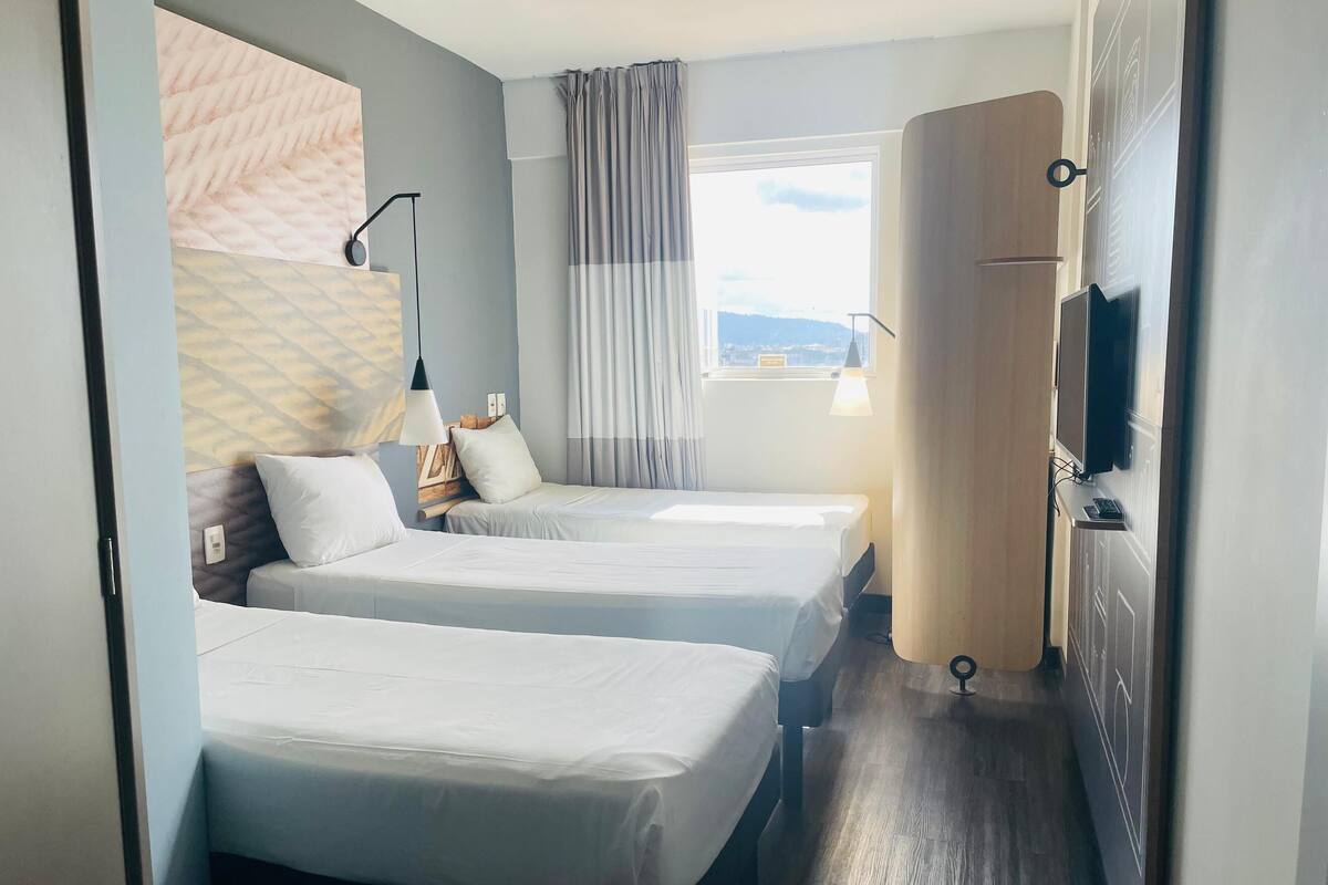 Habitación de hotel con dos camas, una ventana con cortinas, un escritorio con televisor de pantalla plana y televisor montado en la pared.